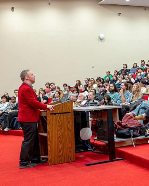 Person wearing a red jacket speaks at a podium, to an auditorium full of students
