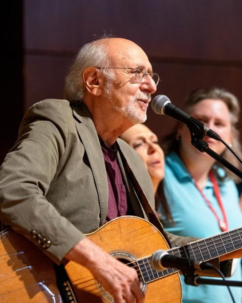 		Peter Yarrow with guitar, tan jacket, white hair just at the back of his head, white beard and glasses in front of a microphone with three women and a child standing next to him
	