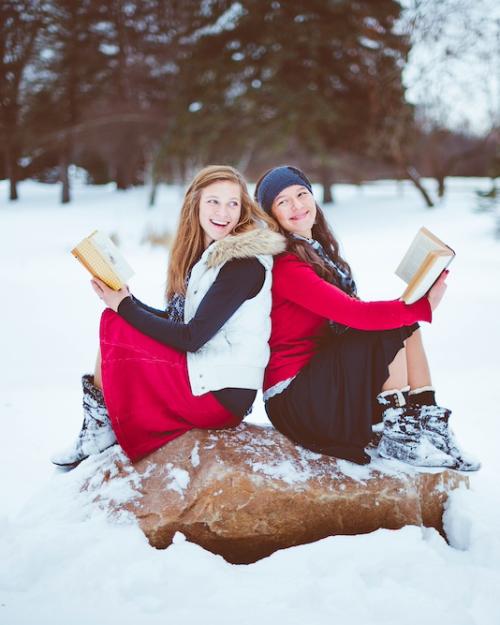 		Two people wearing fashionable red, white and black winter clothing sit back to back on a large rock, each holding a book. They are surrounded by snow
	