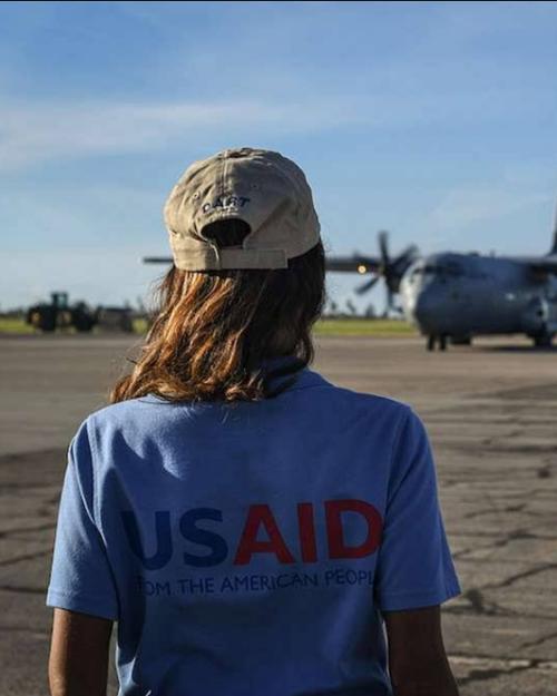 		Two people on a tarmack facing a plane. They're wearing shirts with "USAID" written across the back
	