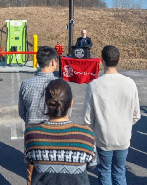 		Sevearl people stand in a parking lot, listening to a speaker who stands at a makeshift podium next to a large green machine
	