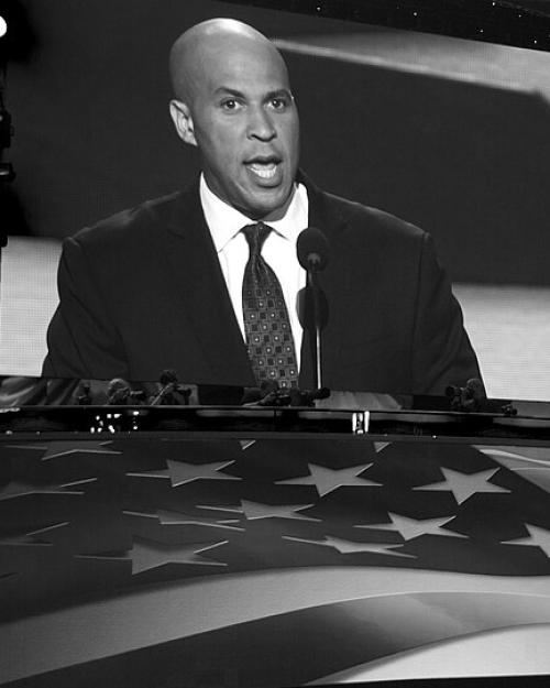 Corey Booker in suit and tie on a large screen with a huge American flag in front of it,