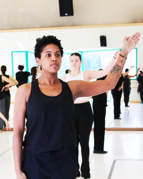 A dancer in the foreground stands upright, holding their left arm at an angle. In the background another dancer replicates the pose, and several other people take different poses or stand around