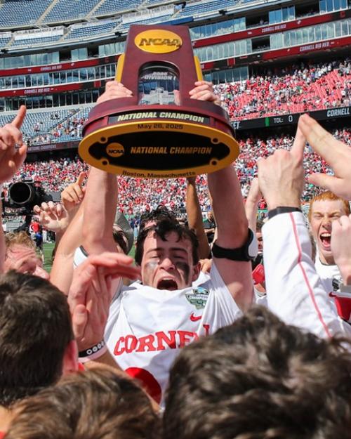A lacross player in white and red uniform holds a trophy aloft; he's surrounded by celebrating teammates