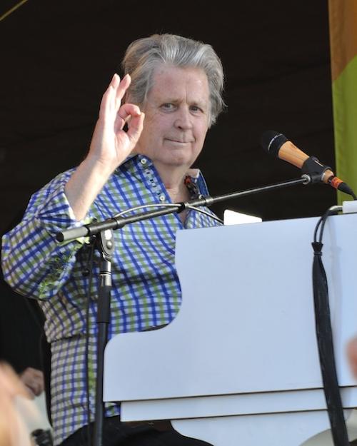 Person sitting at a white piano on a stage, giving an "okay" sign with hand