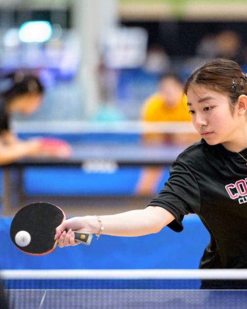 Person in black Cornell t-shirt reaches gracefully to hit a small white ball with a paddle