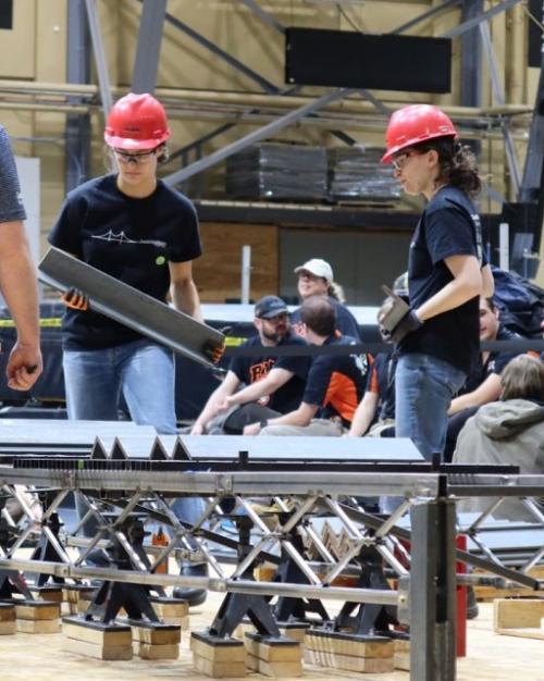 		People in red hard hats construct a structure on the floor of a large indoor space
	