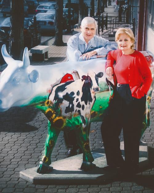 A white haired man and a woman in bright red shirt and sweater stand leaning against a cow with a painting of a cow on it, with a barn in the back and sunflowers and grass on its legs.