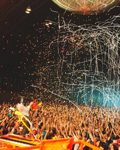 		A crowd raises their hands with excitement as a band plays on a stage (barely in the image) and confetti rains down from above
	