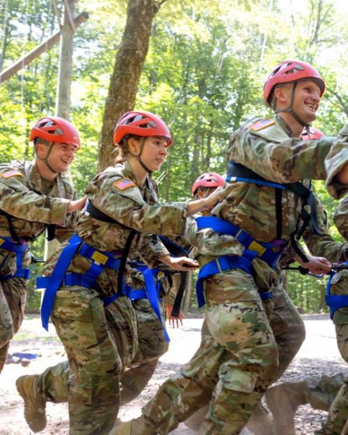 Several people in Army fatigues and red helmets run, strapped together