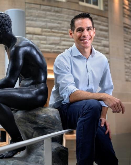 		Person sitting casually back to back with a statue of a seated human figure carved from black stone
	