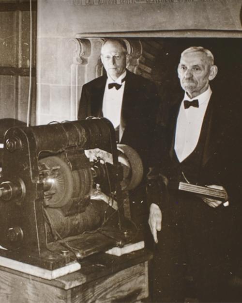 Historic black and white photo of three people in evening coats standing around a machine