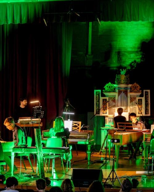 A stage bathed in green light: several people play on keyboard instruments, including standing sythesizers, pianos, and an organ