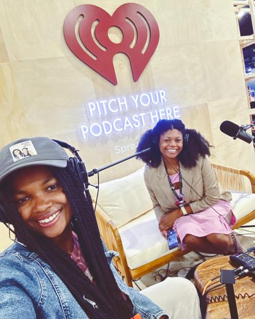 Two people wearing headphones and recording equipment under a neon sign: Pitch your podcast here