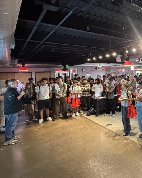 Lots of students standing in a big space with bright lights overhead facing someone with a microphone.