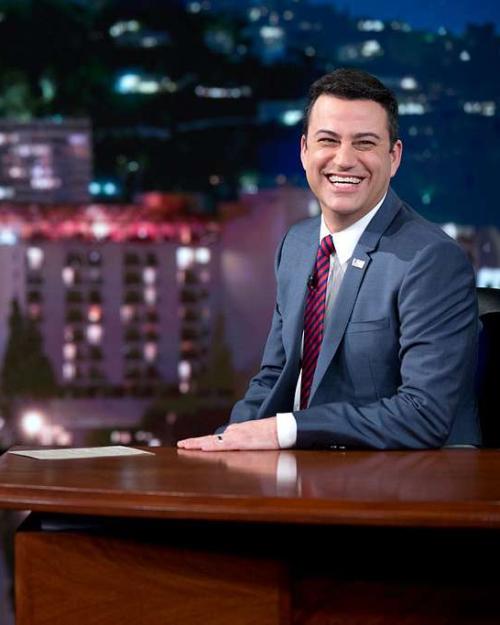 Two people sit at a desk on the set of a late night TV chat show