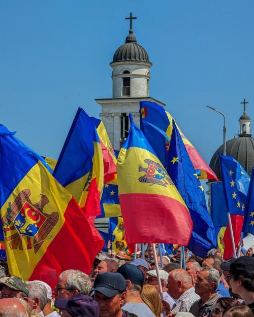 Blue sky, stone spire with a cross on top, people holding red, yellow and blue flags