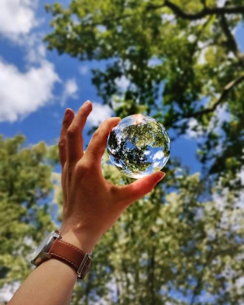 Hand holding a glass sphere up toward the sky so it reflects trees and clouds, making the ball look like a tiny planet Earth