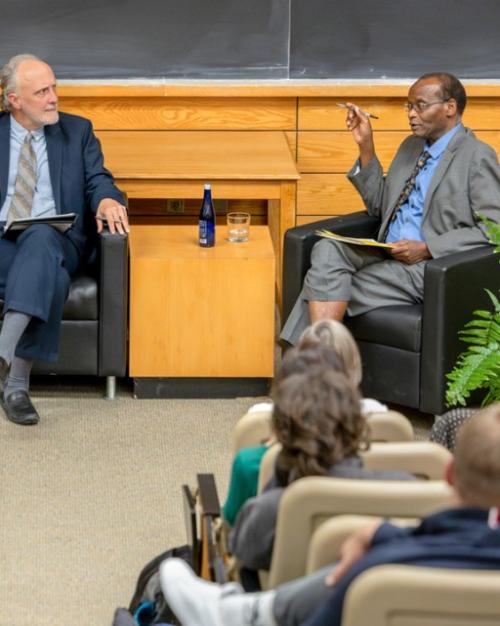 		Three people sit in chairs in front of audience during a debate
	