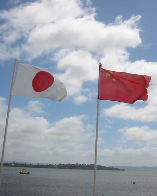		Flags of east Asia on poles near a body of water. Blue sky behind them
	