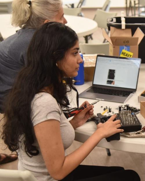 Two people sit at a table loaded with equipment, assembling a robot