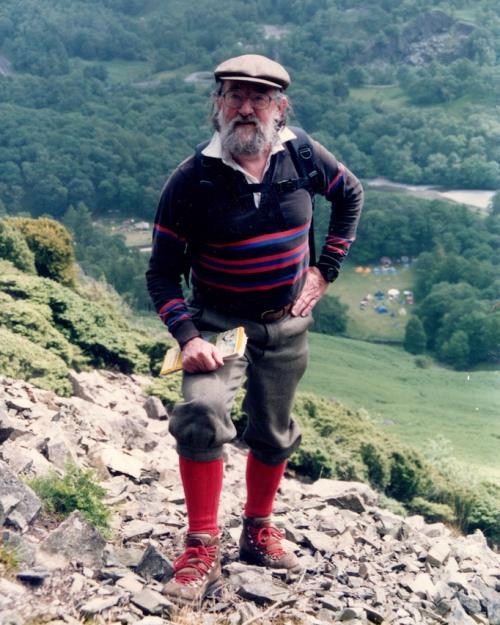 Person wearing a cap, sweater, wool hiking pants and sturdy boots, poses on rocky ground of a very steep hill