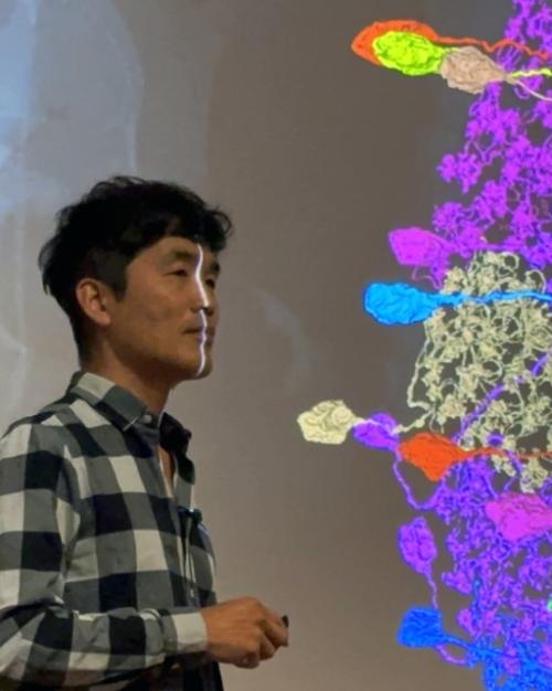 Man with checkered shirt standing in front of a screen with a multi-colored quilt of neurons
