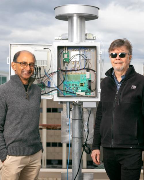 		Three people stand next to a silver device, about a foot taller than the people, made of a post, a box with electronics inside, and a circular top
	