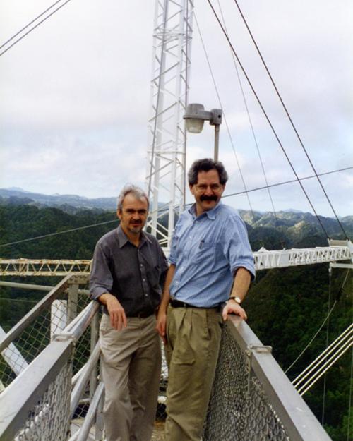 Two people stand on a narrow metal bridge with a crane-like structure rising up behind them