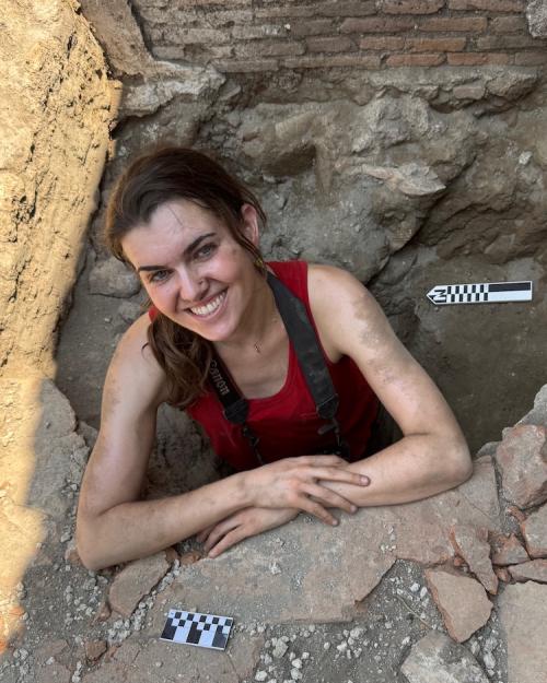 		Person with dust on face and arms, smiling while standing in a hole in the ground, an archaeological dig site
	