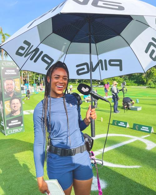 		Person wearing stylish athletic gear, holding a PING umbrella and a microphone, broadcasting at a golf event
	