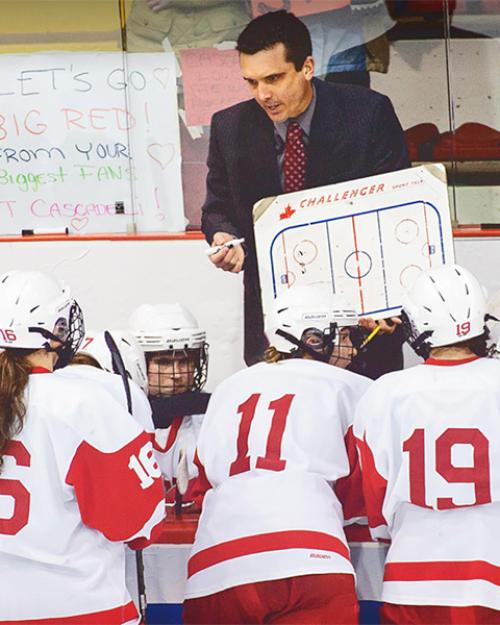 		A coach with a white board in front of several hockey players in red and white jerseys
	