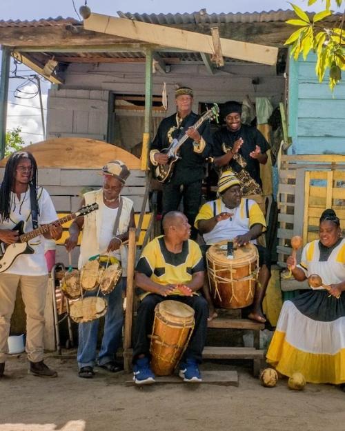 Several people standing around and sitting on porch steps in the shade, playing drums, guitars and maracas