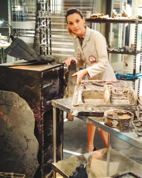 		Actor on the set of a lab, wearing a chic trench coat, among a collection of burned or corrupted objects
	