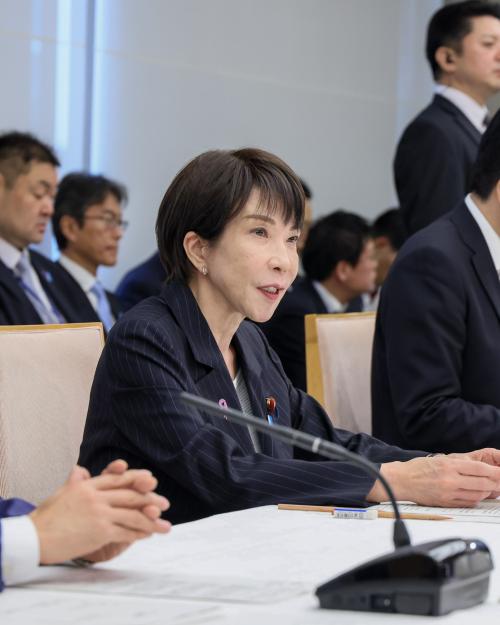 Sanae Takaichi in dark suit jacket sits at a table with microphones between two men, and more men sitting in the room behind her