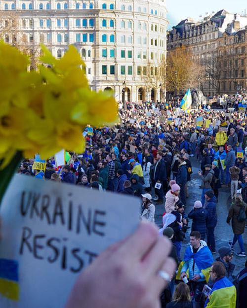 A crowd gathered, holding blue and yellow flags; hands in the foreground hold a sign that says "Ukraine Resist"