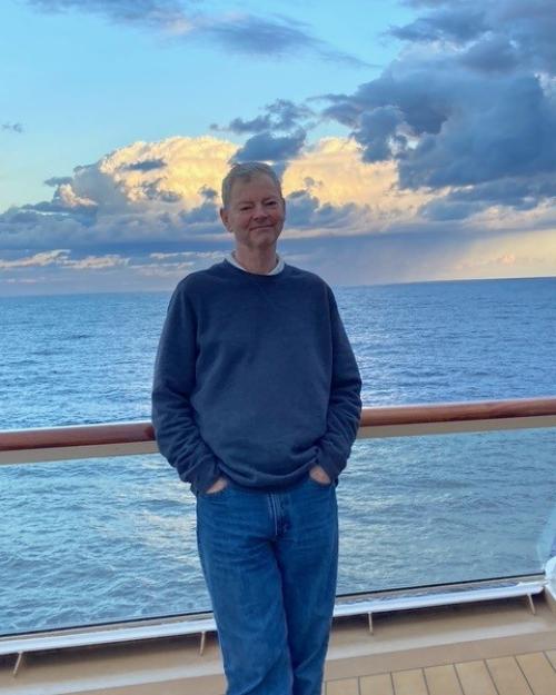 Robert Sullivan, with short gray hair and a closed-mouth smile, leaning against a ship's rail with the sea and clouds behind him.
