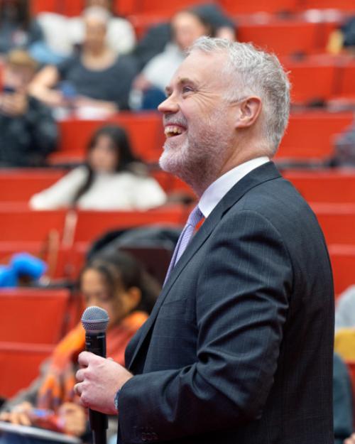 Person in suit holding microphone, in front of a bank of red seats, some occupied