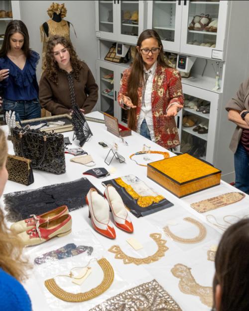 Several people clustered around a large table, on which shoes, handbags, boxes and collars are displayed