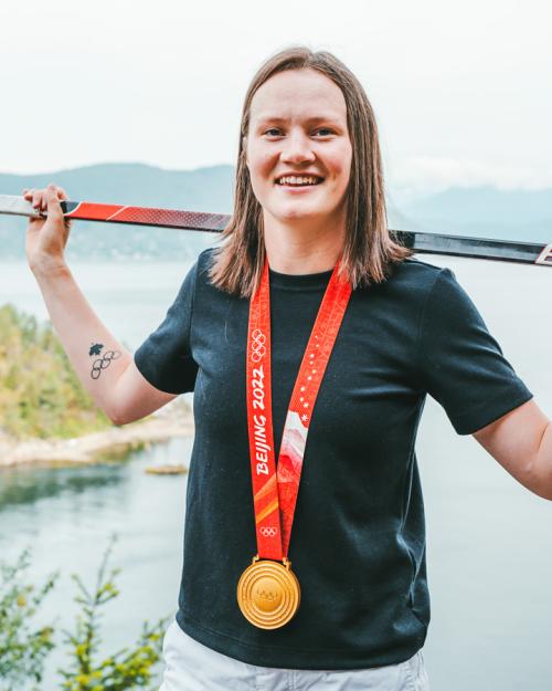 Person standing in front of a body of water, balancing a hockey stick across her back and wearing a gold medal on a red ribbon