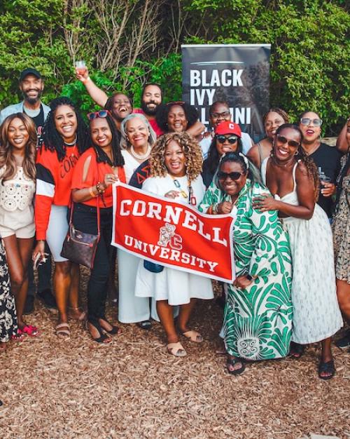 A few dozen people in summer fashions cluster together with a Cornell University banner