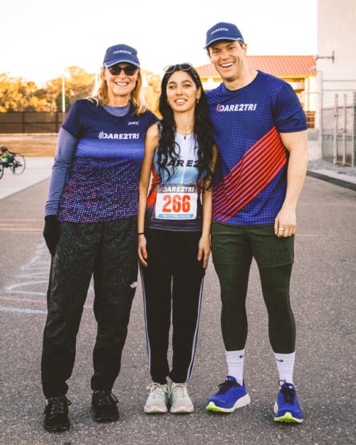 A smiling woman and man in caps and "DARE2TRI" T-shirt stand on either side of a young woman athlete wearing the number "266" on her shirt
