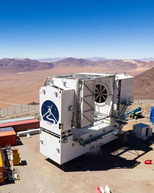 The telescope, a large white structure with the logo on the front, with construction boxes around it and mountain tops and a plateau in the distance.