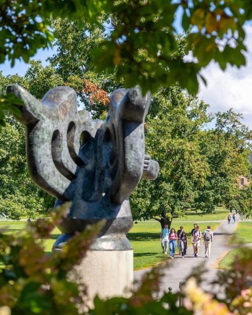 an abstract sculpture in the foreground with a grassy college quadrangle in the distance, bordered by low buildings