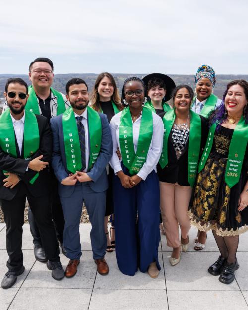 Several people, all wearing green honorary stoles, stand together on a rooftop with a view of a college campus