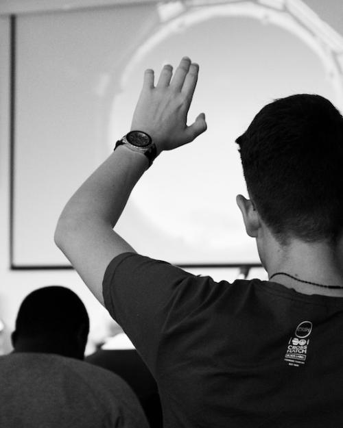 Person seen from the back with hand raised in an audience facing a speaker