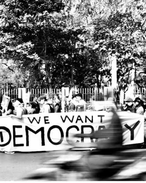 Black and white photo of many people walking together down a street, holding a large banner that reads "We Want Democracy"