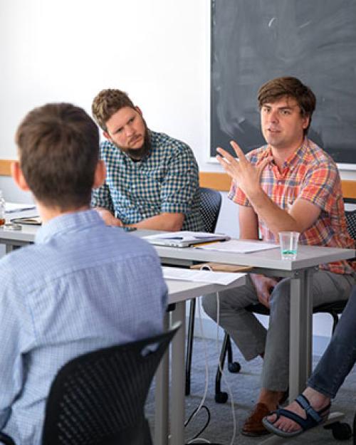 Students in classroom sitting around table discussing a paper.
