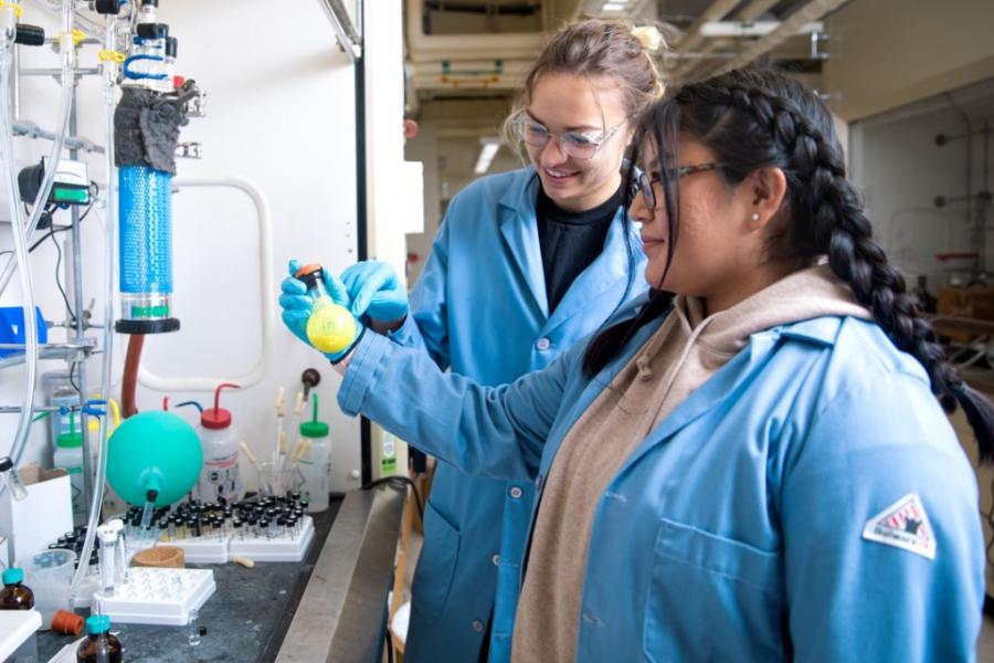 Two students wearing lab coats examining a beaker of something yellow in a lab.