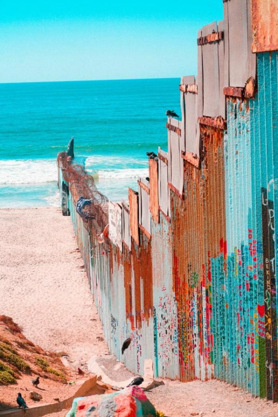 A border wall painted different colors blocking a section of beach with the ocean visible. 